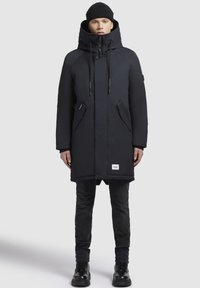 Person med en lång svart huva vinterjacka, svart mössa, svarta byxor och svarta stövlar, som står mot en enkel vit bakgrund.