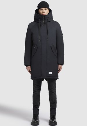 Parka nera isolata con colletto alto, cappuccio regolabile e tasche frontali. Indossata con pantaloni e stivali neri. Presenta un branding minimale.