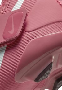 Sportovní bota Nike v růžové barvě, s texturovaným povrchem, nastavitelným páskem a hladkými akcenty. Výrazný design obsahuje vroubkované vzory.