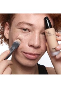 Kiharatukkainen henkilö levittää meikkivoidetta poskelle siveltimellä, pitäen kädessään KIKO Milano Love Fusion 24H Moisture Radiant Foundation -pulloa.