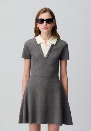 Robe grise à manches courtes avec un décolleté en V et un col en tricot couleur crème. Haut ajusté, jupe évasée, associée à des lunettes de soleil noires.