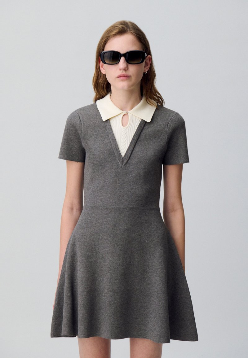 Robe grise à manches courtes avec un décolleté en V et un col en tricot couleur crème. Haut ajusté, jupe évasée, associée à des lunettes de soleil noires.
