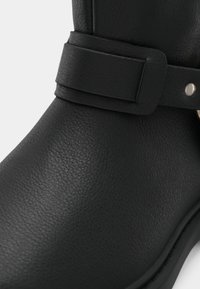 Černý kotník s texturovaným koženým povrchem, širokým popruhem s obdélníkovou sponou a hladkou černou podrážkou.