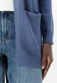 Stickad cardigan i mörkblått, med en enda framficka och ribbade ärmslut. Matchad med ljusblå denimjeans. Slät textur.