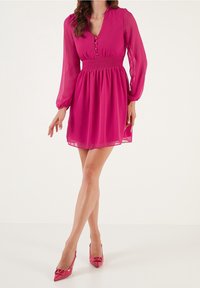 Fuchsia kort klänning med långa genomskinliga ärmar, v-ringning, knapdetails och smockad midja. Matchas med högklackade skor med spetsig tå.