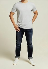 Homme portant un t-shirt gris clair à manches courtes, un jean foncé retroussé et des baskets blanches, debout les mains sur les hanches.