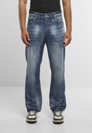 Mężczyzna ubrany w luźne, niebieskie, przetarte dżinsy, czarny top na ramiączkach oraz czarno-białe trampki, stojący na jednolitym szarym tle.