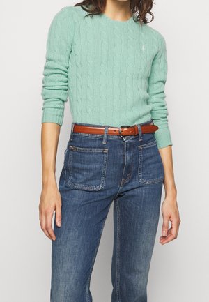 Femme portant un pull en maille torsadée vert menthe à manches longues, rentré dans un jean taille haute bleu avec des poches plaquées à l'avant et une ceinture marron.