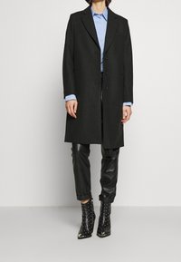 Long black coat made of wool, featuring a notched collar, three buttons, and a straight cut. Worn over a light blue shirt and black leather pants.