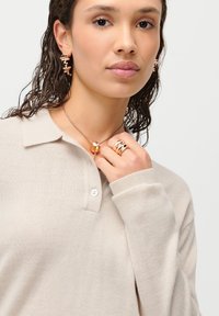 Suéter bege de malha com colarinho e um botão. Acessorizado com um colar dourado em camadas, anéis de ouro e brincos dourados intrincados.