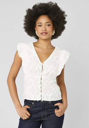 Femme aux cheveux bouclés porte un chemisier en dentelle florale blanche avec des manches à volants et un jean bleu foncé, debout les mains dans les poches.