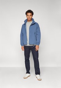 Blue lightweight jacket with a stand-up collar, elastic cuffs, and drawstring hem; layered over a gray sweater and paired with dark jeans.