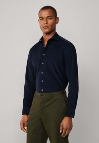 Marineblauw langemouw shirt van zachte stof met knoopsluiting, voorzien van een puntig boord en contrasterende knopen, gecombineerd met olijfgroene broeken.