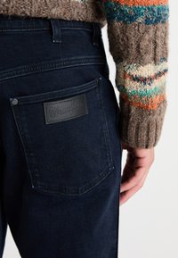 Jeans Wrangler azul oscuro con un parche de cuero negro en el bolsillo trasero, con costuras tonales y un primer plano de un suéter texturizado.