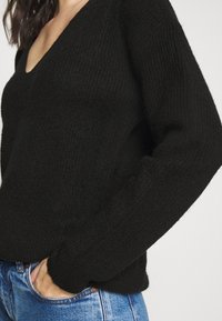 Svart stickad tröja med V-hals, med ett texturerat tyg och ribbade ärmslut. Matchas med hög midja blå denimjeans.