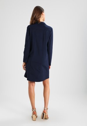 Vrouw die een marineblauwe jurk met lange mouwen en gouden sandalen met hoge hakken draagt, staand met haar rug naar een eenvoudige lichte achtergrond.