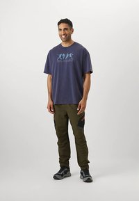 Tamsiai mėlyna medvilninė marškinėliai su grafiniu atspaudimu, derinami su alyvuogių žaliais krovinių kelnėmis su juodais akcentais ir keliais kišenėmis.