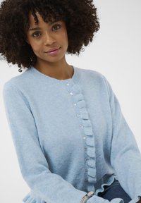 Világoskék kötött kardigán fodros elejével és kerek nyakvágásával, kerek gombokkal díszítve a záródás mentén.