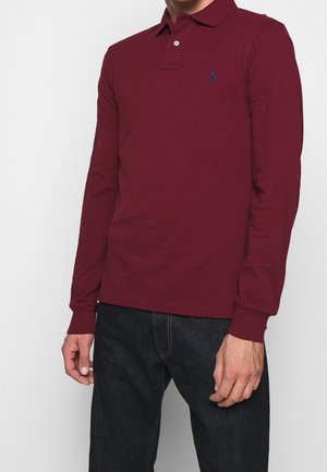 Homme portant un polo à manches longues marron rouge avec un petit logo bleu sur la poitrine et un jean bleu foncé sur un fond blanc uni.