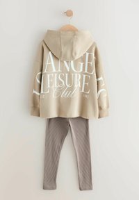 Beige hoodie med vit text, "Los Angeles Leisure Club," i kombination med ljusbruna ribbade leggings. Hoodien har snörning och känguruficka.