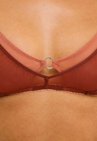 Bra i rostfärg med genomskinlig mesh-tyg, V-formad design och en central cirkulär accent. Inkluderar delikata vågiga kanter.