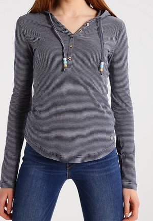 Femme portant un sweat à capuche à manches longues rayé bleu marine et blanc avec des cordons ornés de perles en bois, associé à un jean bleu foncé.