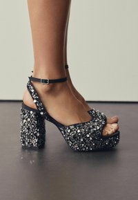 Feet wearing black platform heels with ankle straps, covered in shiny silver sequins, standing on a smooth dark floor.