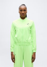 Μια γυναίκα στέκεται με το σώμα στραμμένο προς τα εμπρός, φορώντας ένα φούτερ με φερμουάρ σε νέον πράσινο χρώμα και αντίστοιχο παντελόνι με αθλητικό λογότυπο στο στήθος και στο μηρό.