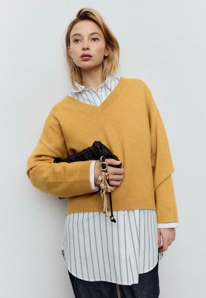 Sinapinkeltainen v-kaula-aukkoinen paita, pehmeää neulosta, yhdistetty valkoiseen raidalliseen paitaan. Pitelee mustaa, teksturoitua clutch-laukkua, jossa koristeelliset metalliosat.