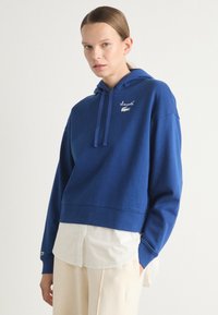 Felpa blu tagliata con cappuccio e coulisse, caratterizzata da un logo ricamato bianco. Indossata sopra una camicia bianca in una vestibilità rilassata. Texture del tessuto morbida.