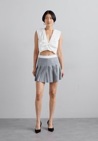 Biały crop top z kokardą; szara plisowana mini spódniczka. Gładki materiał, dopasowany krój i wysoki stan. Czarne szpilki.