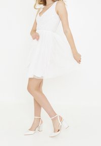 Valkoinen mekko, jossa on paljeteista valmistettu vartalo-osa, V-aukko ja kerroksellinen tyllihame. Yhdistetty valkoisiin lakattuihin kärkitossuihin, joissa on nilkkahihnat.