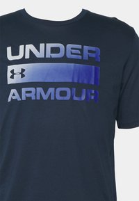 Marineblå bomulls-T-skjorte med iøynefallende "UNDER ARMOUR"-tekst i gradientblå. Korte ermer og rund hals.