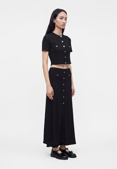 Γυναίκα στέκεται φορώντας μαύρο crop top και μακρύ φούστα με λεπτομέρειες κουμπιών, σε συνδυασμό με μαύρα παπούτσια Mary Jane σε απλό φόντο.