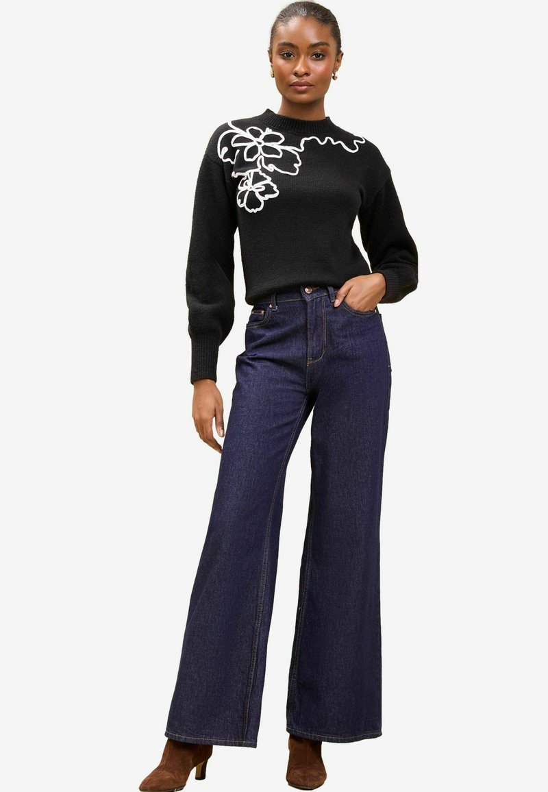 Maglione nero con ricami floreali bianchi, abbinato a jeans a vita alta di jeans blu a gamba larga e stivaletti con tacco marroni.