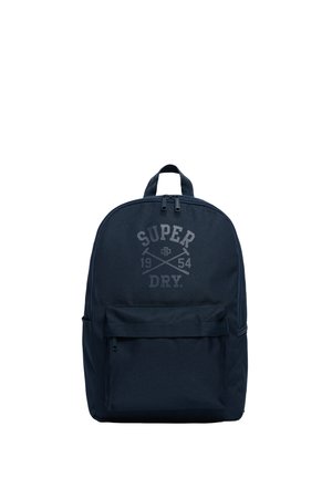 Marineblauer Rucksack mit einer vorderen Reißverschlusstasche, einem oberen Griff und dem in Grau auf dem Hauptfach aufgedruckten "Super Dry 1954"-Logo.