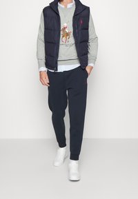 Smanicato imbottito blu navy sopra una felpa grigia con logo polo. Camicia celeste chiara con colletto sotto, abbinata a pantaloni da tuta scuri e sneakers bianche.