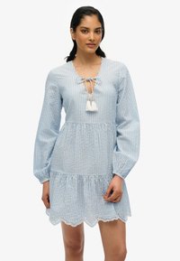 Vestido a rayas azul y blanco con mangas largas, un escote en forma de barco y acentos de borlas. Presenta un dobladillo de volantes en capas con bordes en forma de onda.