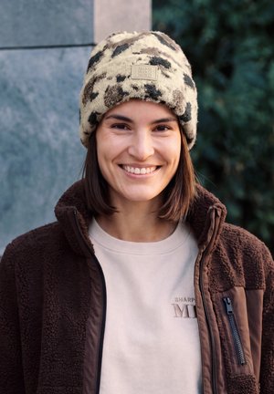 Mujer sonriente al aire libre, con un gorro de punto estampado en crema y negro, una chaqueta polar marrón y una sudadera clara.