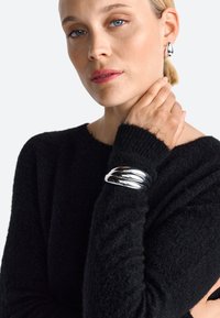 Silberner metallischer Armreif mit drei miteinander verwobenen Bändern, getragen auf einem schwarzen strukturierten Pullover. Die Haut des Models ist hell und das Haar ist blond.