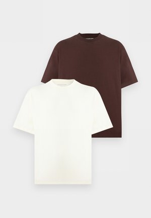 Dwie osoby stoją przy białej ścianie w oversize'owych ciemnobrązowych t-shirtach, czarnych szerokich spodniach i białych trampkach, z rękami w kieszeniach.