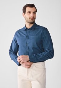 Camisa de botones azul oscuro en tejido suave, con un bolsillo tipo parche, cuello abotonado y mangas largas, combinada con pantalones beige claros.