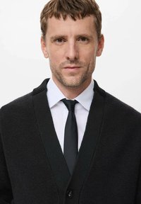 Czarny kardigan z dekoltem w kształcie litery V, teksturowany knit, z zapięciem na guziki, noszony na białej koszuli i czarnym krawacie. Prosty, formalny wygląd.