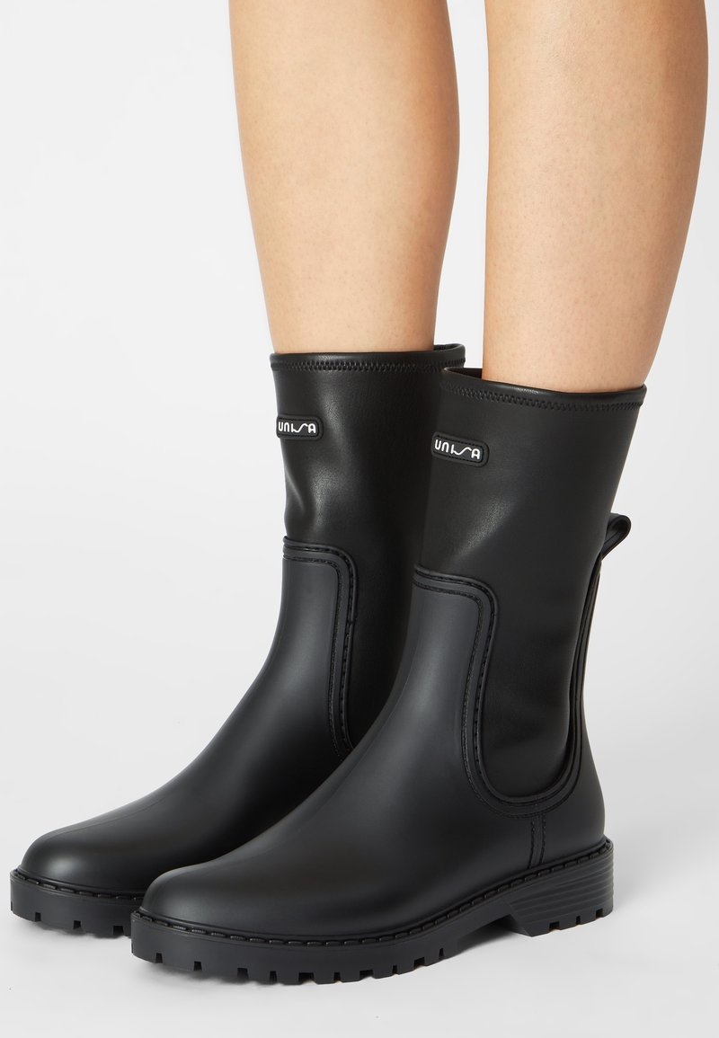 Bottes en caoutchouc noires mi-mollet avec languette de tirage et semelle texturée portées sur des jambes nues, avec un petit logo de marque près du sommet.