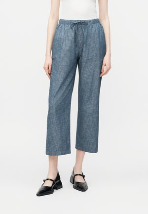 Γυναίκα που φοράει μπλε παντελόνι φαρδύ τύπου cropped με ελαστική μέση και κορδόνι, λευκό τοπ και μαύρα παπούτσια Mary Jane με τακούνι block.