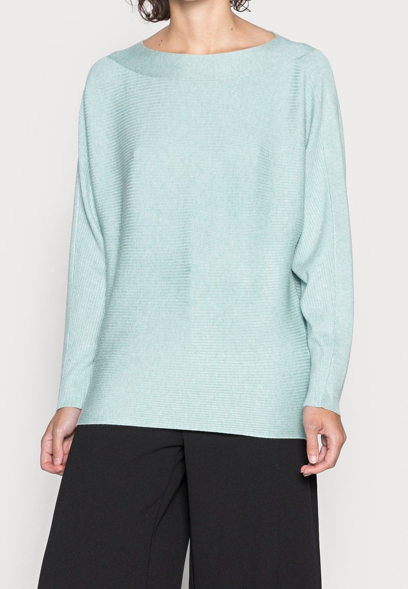Helles, teal-farbenes, lässig geschnittenes Sweatshirt mit weitem Ausschnitt, gerippter Textur und langen Ärmeln, kombiniert mit schwarzen weit geschnittenen Hosen.