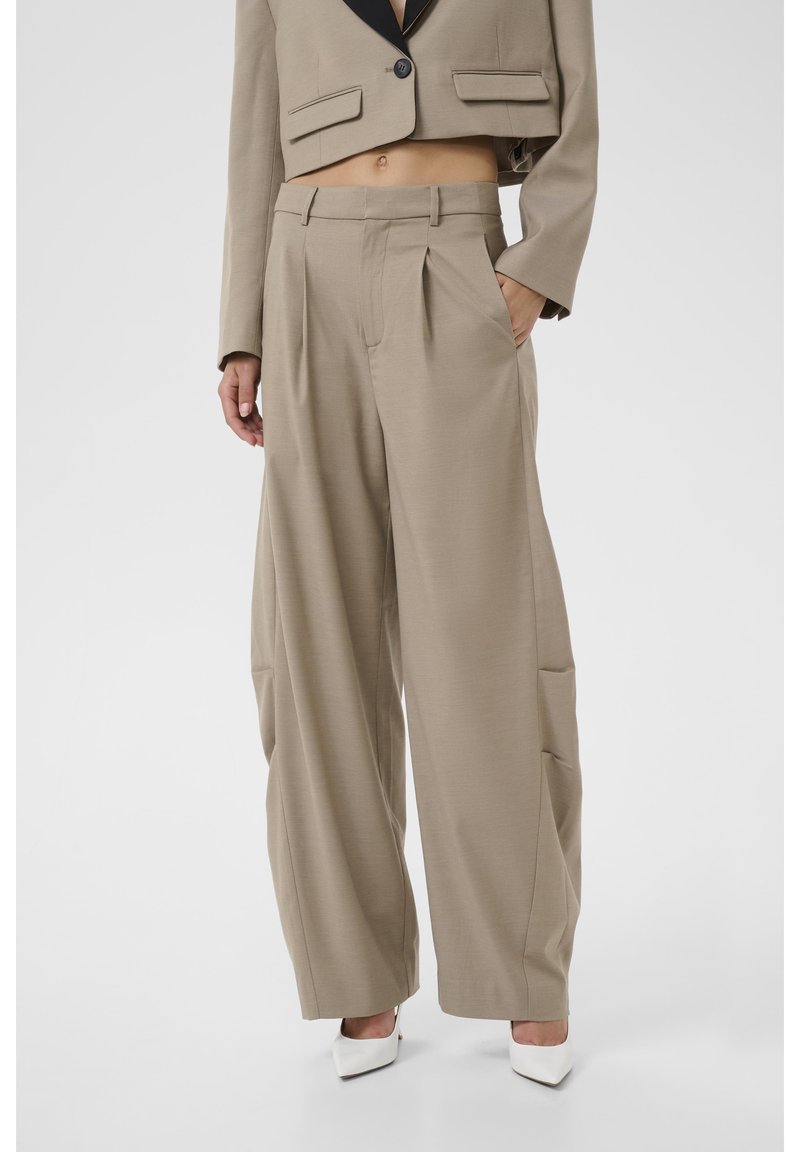 Pantalon beige à jambes larges avec plis porté avec un blazer beige court à revers noirs et talons blancs pointus.
