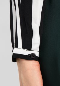 Poignet de chemisier à rayures noires et blanches avec un détail de bouton, fabriqué en tissu lisse, sur un fond vert riche.