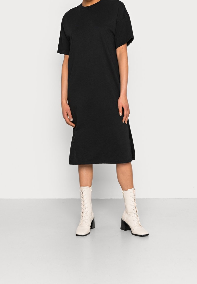 Personne portant une robe midi noire à manches courtes associée à des bottes blanches à lacets mi-mollet à talons, debout devant un fond blanc uni.