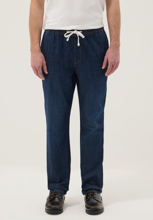 Man met donkerblauwe denim broek met elastische taille en wit trekkoord, en zwarte veterschoenen, tegen een effen achtergrond.